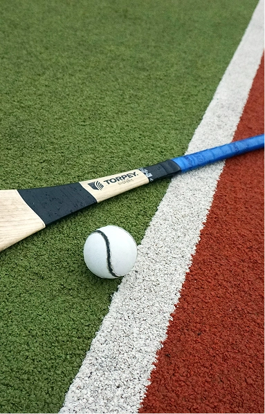 RE4ORM synthetic turf recycling &ndash; A field hockey stick and a white ball with a black stripe rest on green artificial turf, illustrating how the RE4ORM Process can enhance sports surfaces, next to a white line and a red section of the pitch.