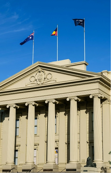 RE4ORM synthetic turf recycling &ndash; A neoclassical building with tall columns and three flags flying on top&mdash;showcasing the RE4ORM Process&mdash;features the Australian flag, the Aboriginal flag, and another unidentified flag, all set against a clear blue sky.