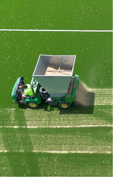 RE4ORM synthetic turf recycling &ndash; A worker drives a green tractor spreading sand or material over artificial turf using the RE4ORM Process, creating visible light stripes across the bright green surface, with a white line in the background.