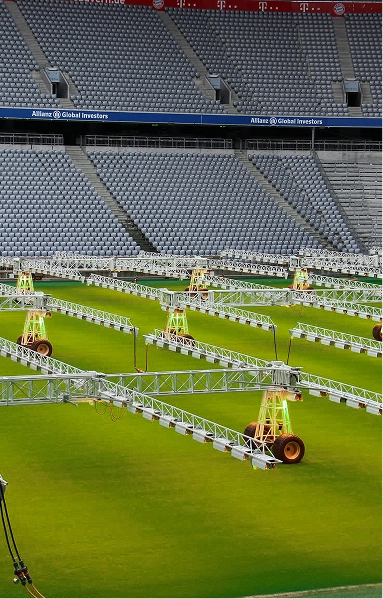 RE4ORM synthetic turf recycling &ndash; Several large light rigs on wheels are positioned across a bright green football pitch in an empty stadium with grey seats. The scene highlights the RE4ORM Process, visible alongside signs for Allianz Global Investors.