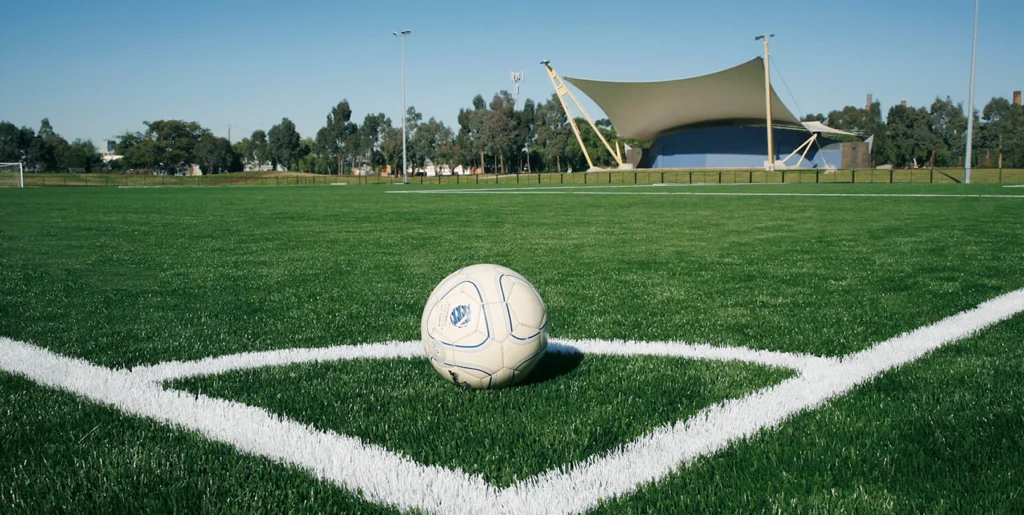 RE4ORM synthetic turf recycling &ndash; A white football rests on the green grass near a corner of a football pitch, with white boundary lines and a large canopy structure and trees in the background under a clear blue sky.