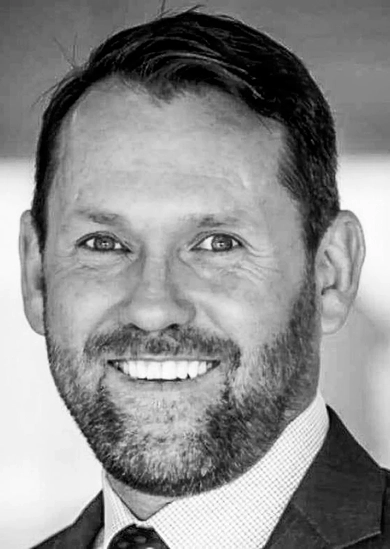 RE4ORM synthetic turf recycling – A man with short dark hair and a beard smiles at the camera. He is wearing a suit and tie. The image is in black and white.