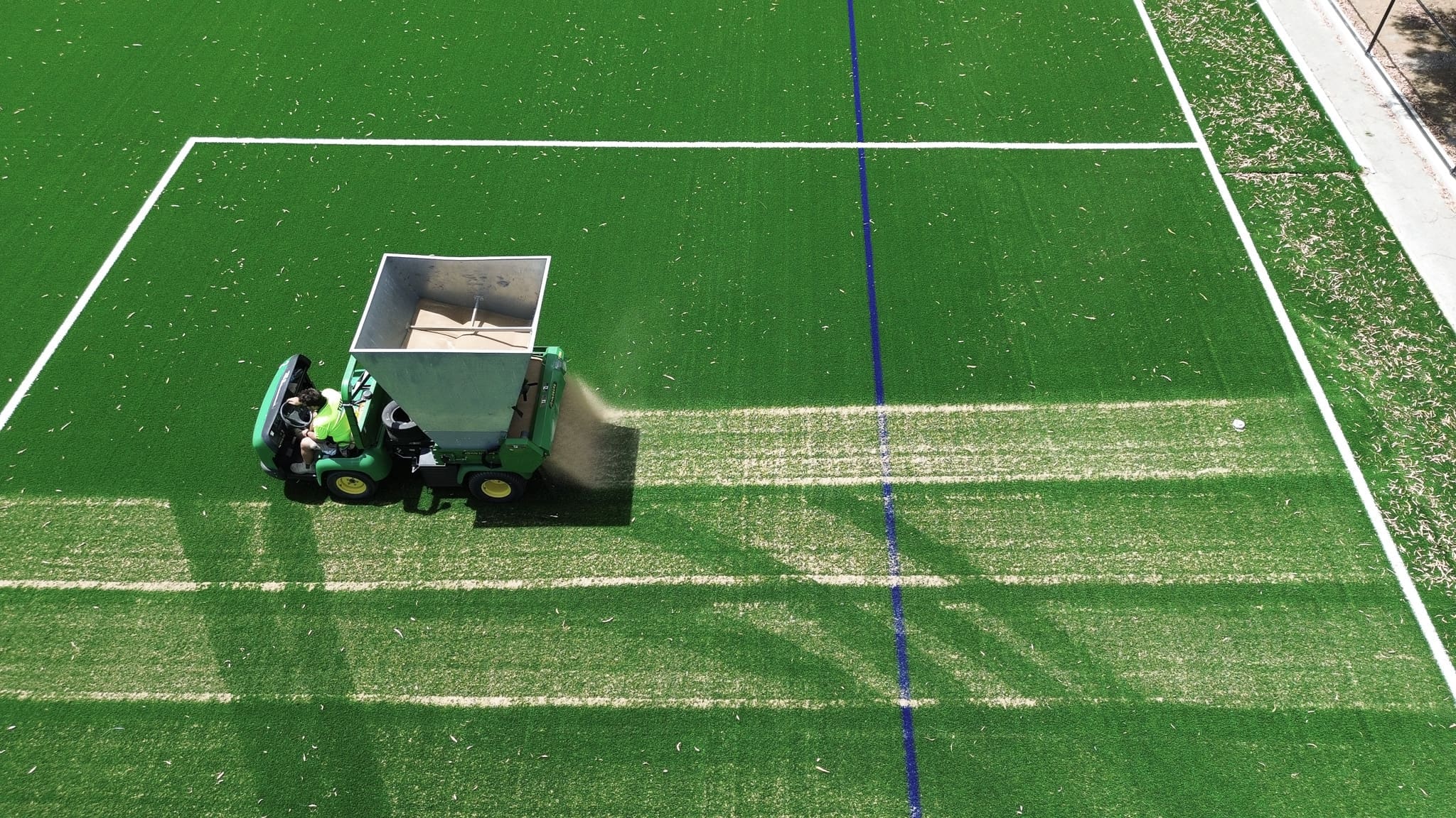RE4ORM synthetic turf recycling – A green maintenance vehicle moves across the artificial turf pitch at Rowville Secondary College, leaving a lighter path behind. White and blue pitch lines are visible, and the scene is viewed from above.
