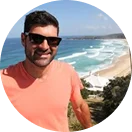 RE4ORM synthetic turf recycling – A man wearing sunglasses and a coral T-shirt smiles at the camera with Kareela Oval and ocean waves in the background on a sunny day.