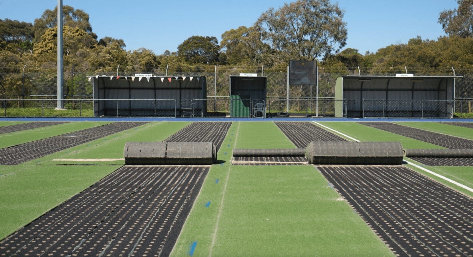 RE4ORM synthetic turf recycling – Rolls of artificial turf and black rubber mats are laid out on the Kingston Heath Reserve sports pitch, with benches and trees in the background, suggesting pitch renovation or maintenance work is underway.
