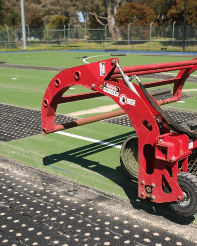 RE4ORM synthetic turf recycling – A close-up of a red piece of construction equipment with a claw-like attachment on the artificial turf pitch at Kingston Heath Reserve, with trees and a fence in the background.