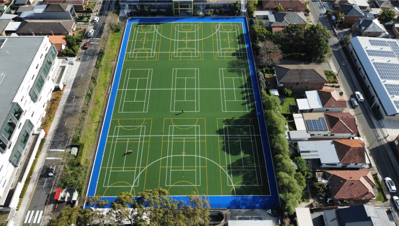 RE4ORM synthetic turf recycling – Aerial view of a large, green multi-sport court with bright blue borders, surrounded by residential houses and streets; a few people are visible on the pitch.