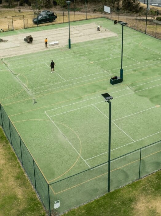 RE4ORM synthetic turf recycling &ndash; Aerial view of a green tennis court with two people; one stands near the net, and the other near the baseline. A car and equipment are visible outside the fenced court, where the RE4ORM method is applied to rejuvenate its slightly worn surface.