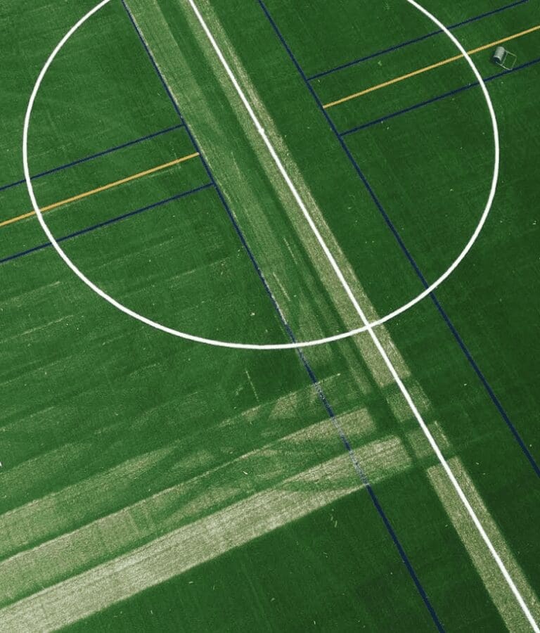 RE4ORM synthetic turf recycling – Aerial view of a green sports pitch at Rowville Secondary College, showing a white centre circle and multiple intersecting pitch lines in white, yellow, and blue. Some areas of the grass appear worn or marked.