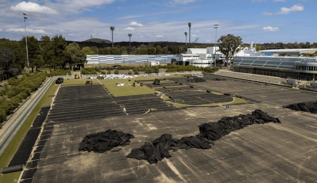 RE4ORM synthetic turf recycling &ndash; A large outdoor sports pitch at an Australian sports institute is under construction, with sections of artificial turf removed and black materials and rolls laid out. Buildings, trees, and stadium floodlights can be seen in the background beneath a partly cloudy sky.