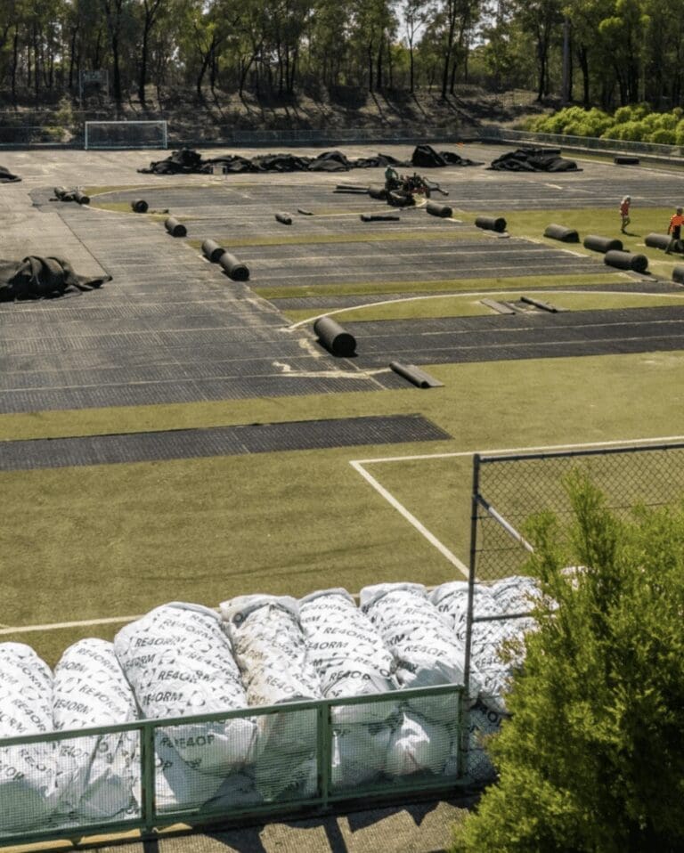 RE4ORM synthetic turf recycling – Workers install artificial turf on a football pitch at an Australian sports institute. Rolled-up turf and construction materials are scattered across the pitch, with several white bags in the foreground. Trees and a goalpost stand in the background.
