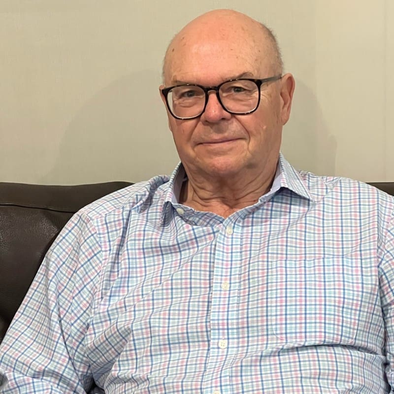 RE4ORM synthetic turf recycling – An older man with glasses, wearing a light-coloured, checked shirt sits on a dark sofa, looking at the camera with a neutral expression against a plain background.