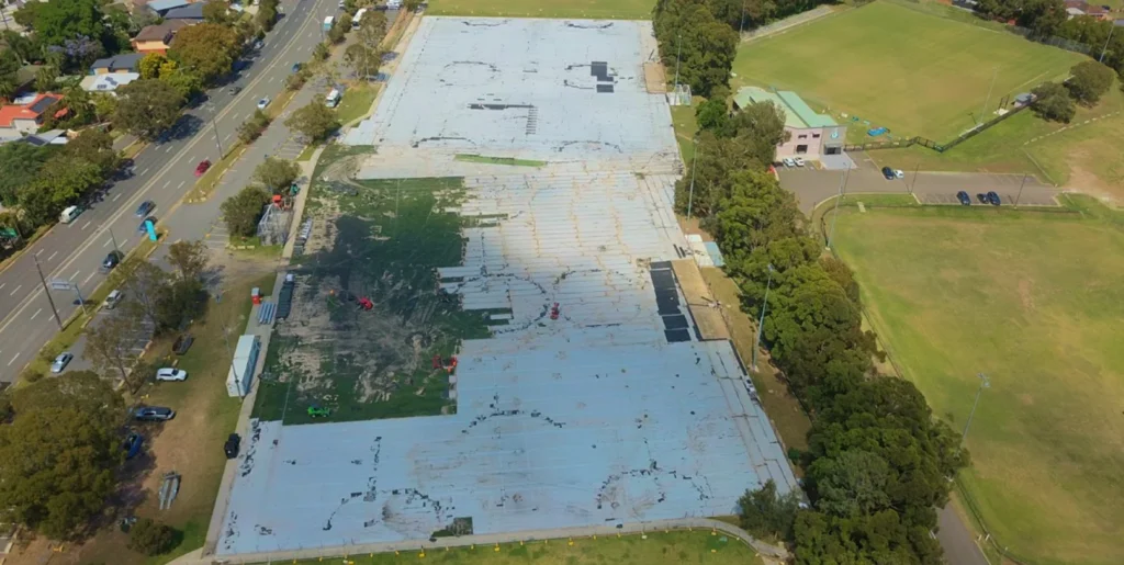 RE4ORM synthetic turf recycling &ndash; Aerial view of a large, mostly empty concrete car park near Kareela Oval, with patches of greenery and equipment, surrounded by trees, grass fields, and a road lined with houses on the left side.