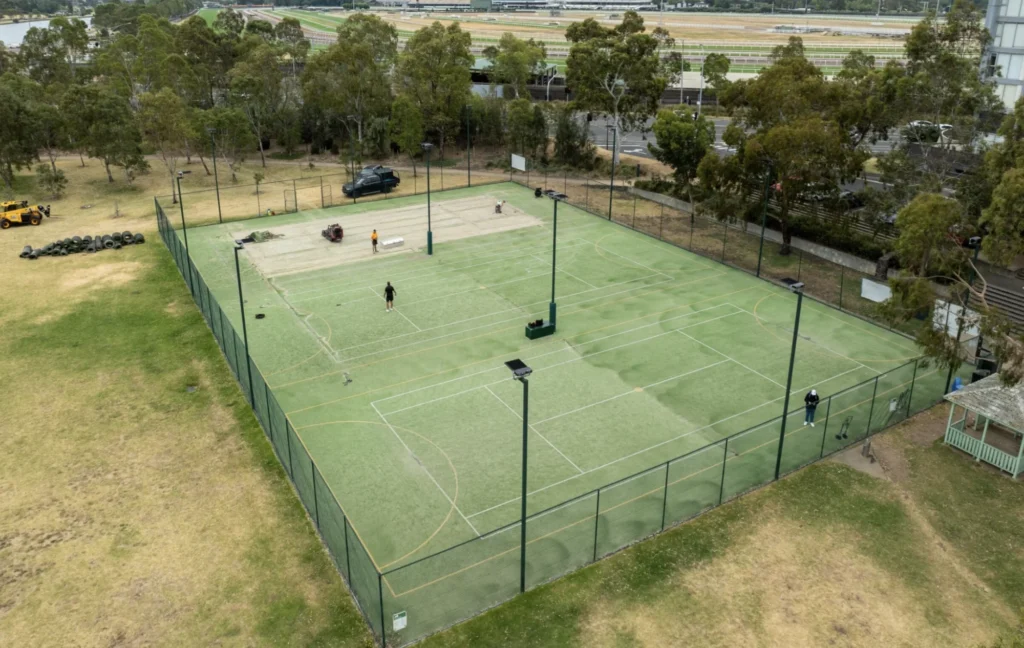 RE4ORM synthetic turf recycling &ndash; Aerial view of a fenced outdoor sports court in Kensington Banks, marked for multiple games, surrounded by grass and trees. A few people are on the court, with some sports equipment visible in the area.